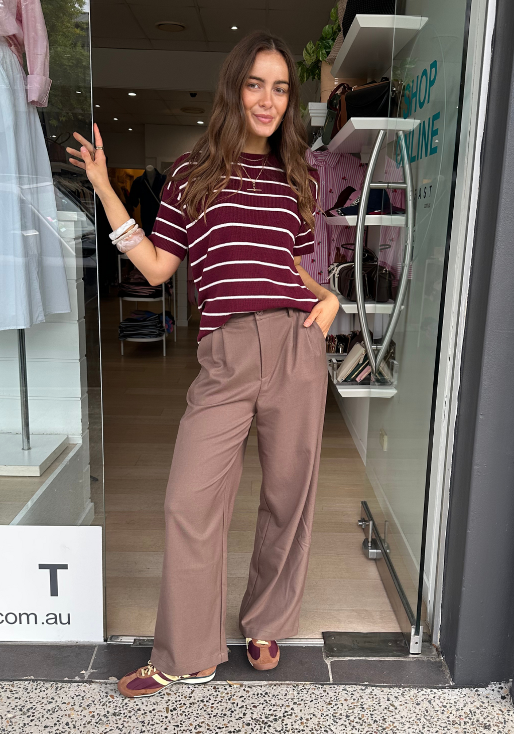 Woman in a striped shirt and brown pants standing outside a store entrance.